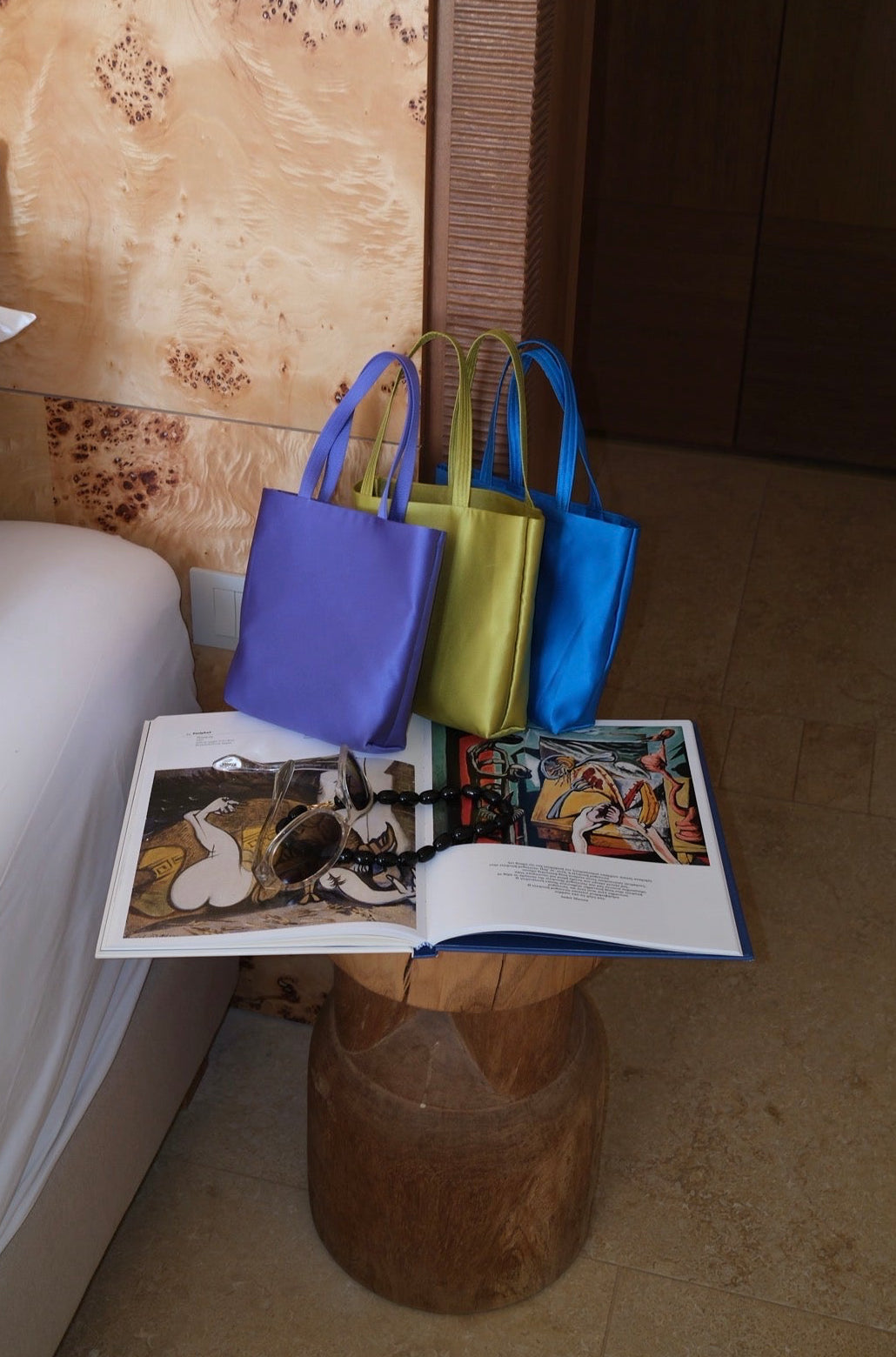 Three colorful mini satin tote bags in purple, yellow-green, and blue are arranged on a wooden stool next to a bed, with an open magazine in front of them.